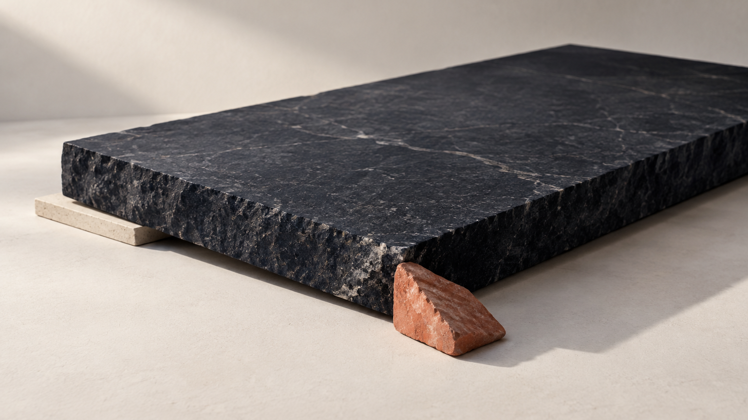 A wide-angle studio still life on a pale grey sweep: one thick slab of dark stone lies flat, a single thin pale board is wedged beneath it as a supporting shim, and a small angular wedge rests against the slab's lower edge touching only part of it. The diagonal camera axis runs from lower left to upper right so the wedge clearly bites one corner while the slab stays level and unbroken. No text, no letters, no numbers, no symbols, no logos, no people, no hands.