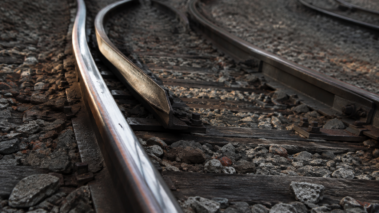 A close, low-angle oblique view of a real railway junction where one running rail splits away and continues into three separate diverging routes across ballast. The chosen route is rendered in sharp focus with polished worn steel, while the other diverging branches sit in softer focus with duller weathered metal, so the eye reads one path taken and other branches still to be rechecked. Textures of gravel, sleeper grain and oiled steel dominate. No signs, no text, no labels, no numbers, no paper.